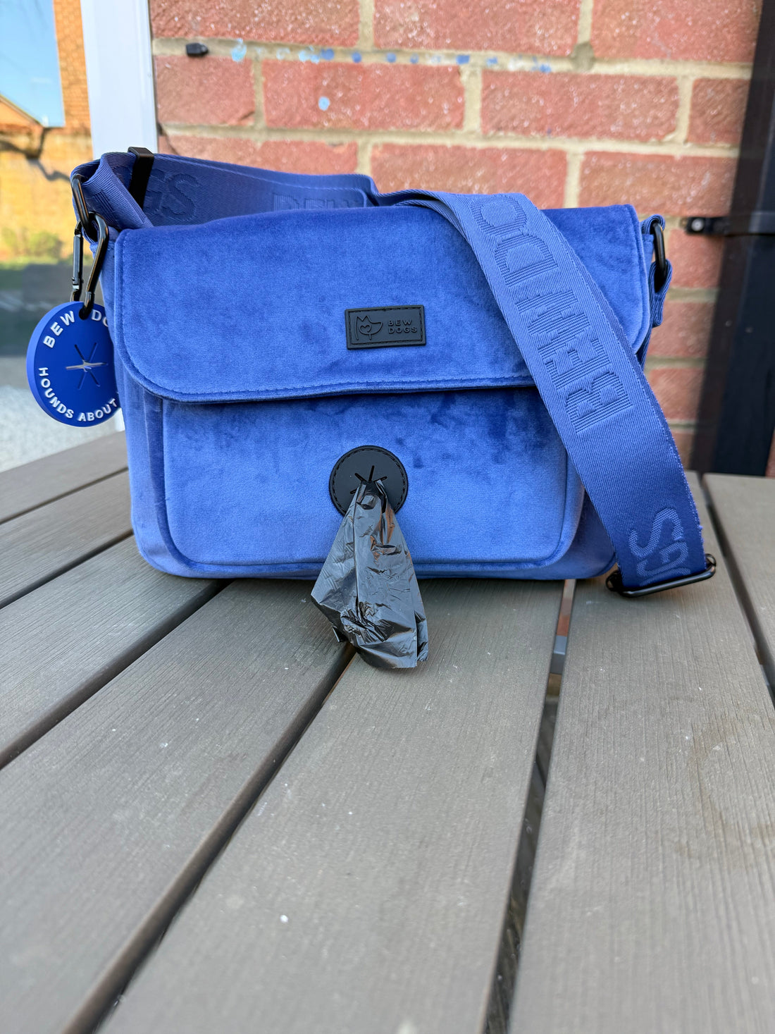 A blue velvet bew dogs walking bag, with poop loop & special compartments, sits in front of a brick wall. 