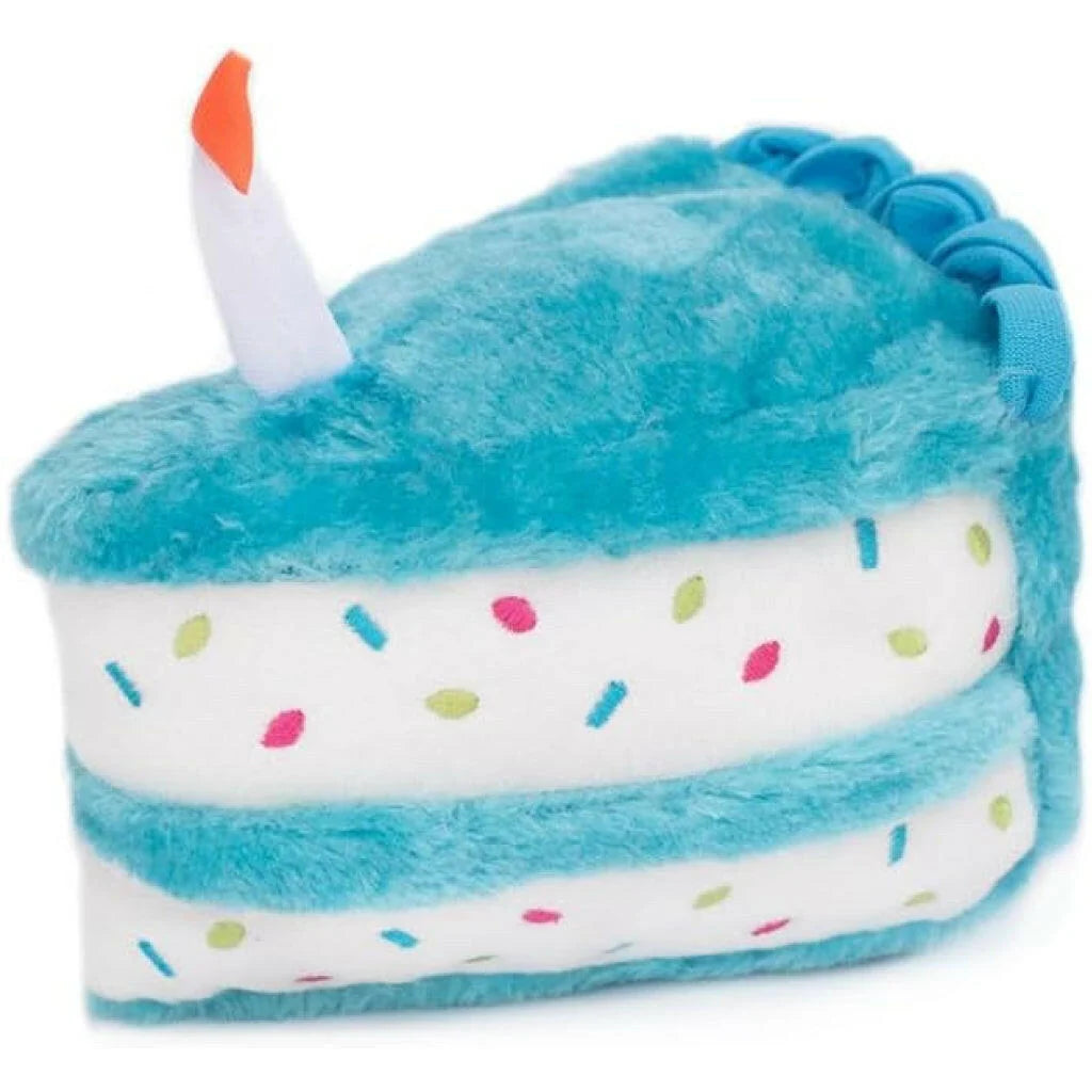 A plush dog toy shaped like a slice of cake with turquoise blue frosting, white layers, colorful sprinkle details, and a white candle with an orange flame on top, shown on a plain white background.