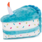 A plush dog toy shaped like a slice of cake with turquoise blue frosting, white layers, colorful sprinkle details, and a white candle with an orange flame on top, shown on a plain white background.