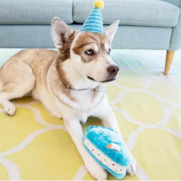 A husky dog wearing a blue party hat lies on a yellow patterned rug, holding a blue plush cake-shaped toy in its paws, the perfect birthday gift for all dogs 