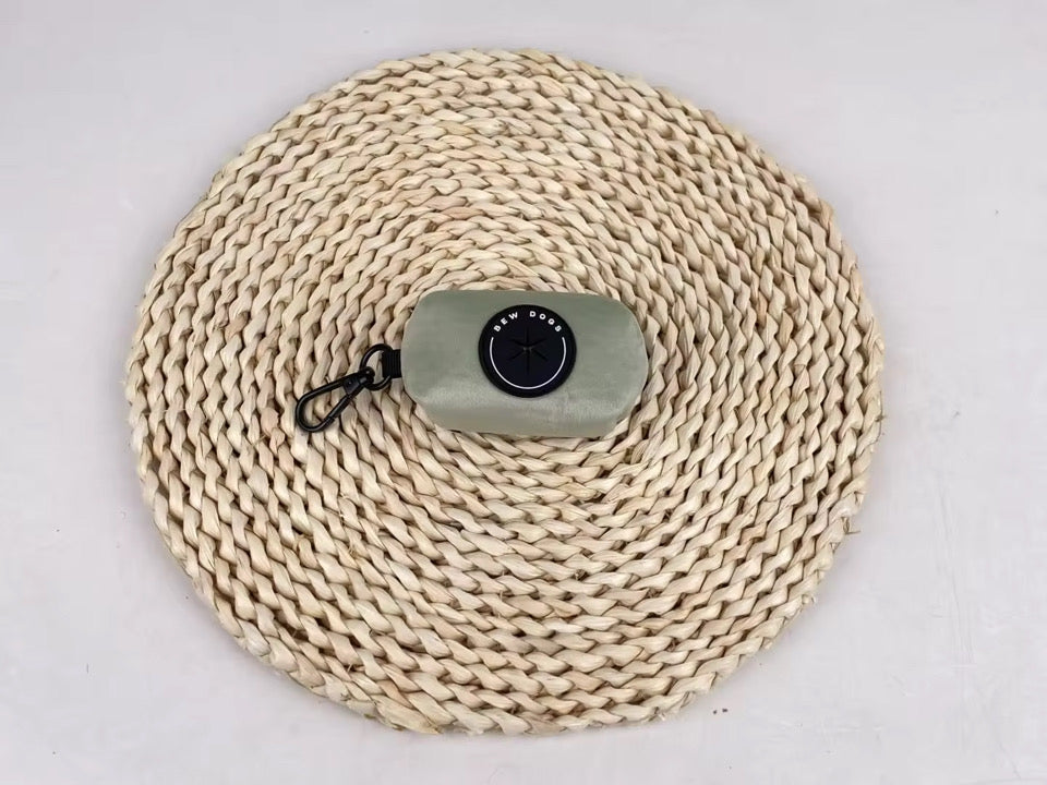 Sundown Collection dog poo bag holder in sage green, sits on a woven mat against a grey background