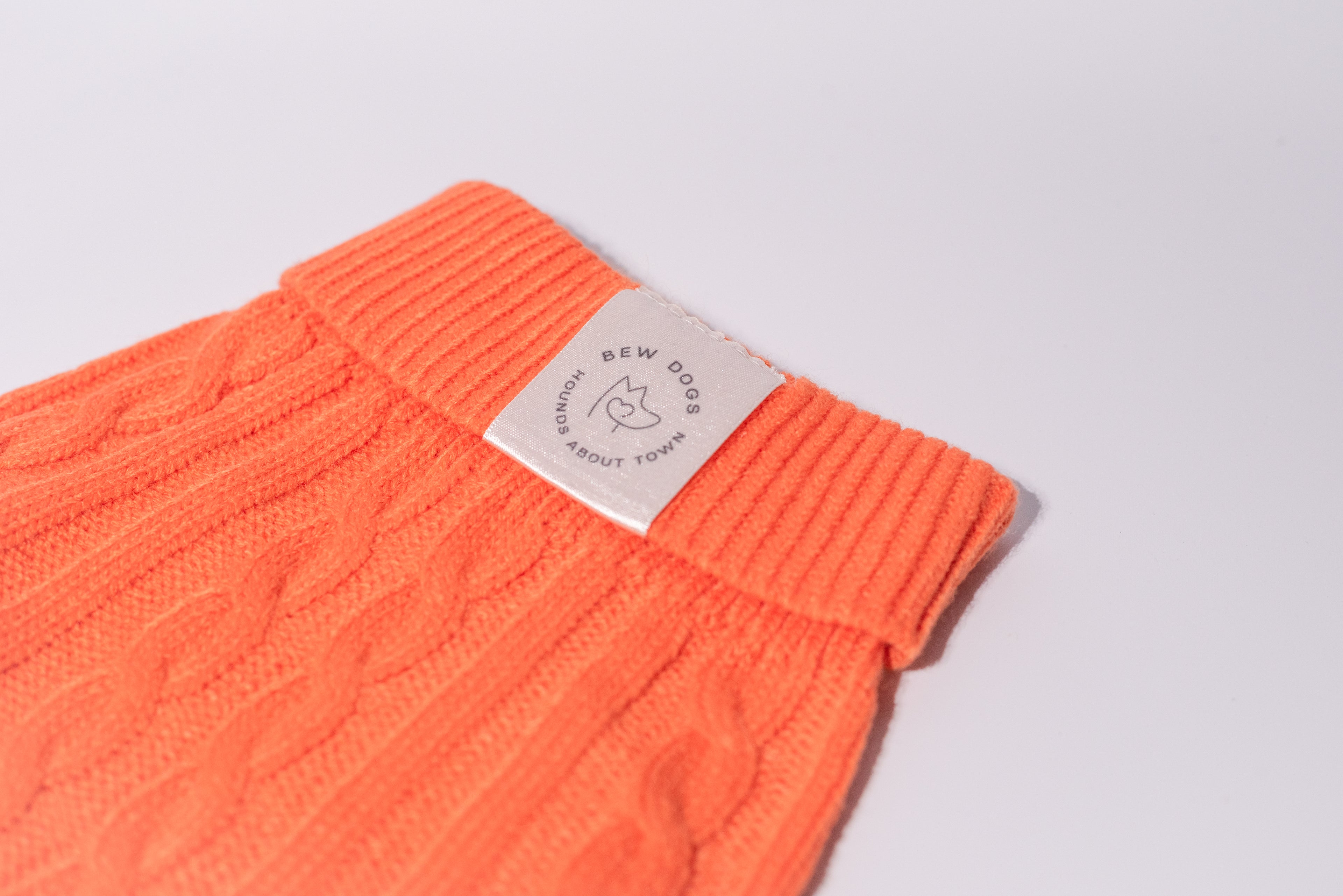 A close-up of a bright orange cable-knit dog sweater with a ribbed cuff, featuring a white fabric brand tag that reads 'Bew Dogs'.