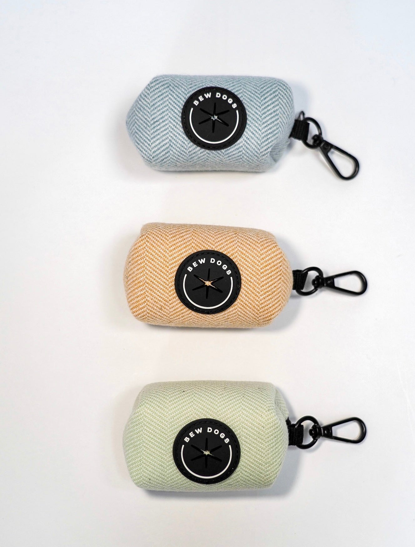 three herringbone holders to keep poo bags handy on dog walks, by attaching to a lead or bag, are shown in blue, cream and green - set against a white background