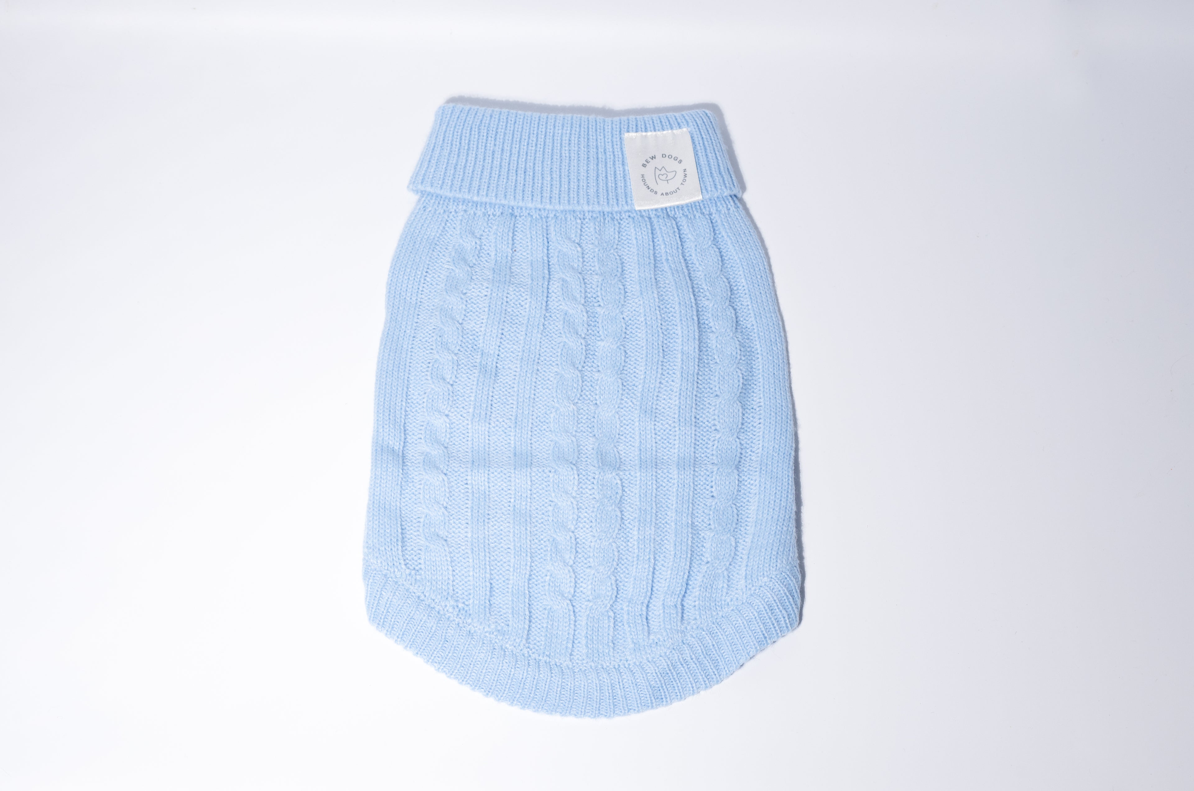 A light blue cable-knit dog sweater with a ribbed collar and a small white Bew Dogs brand patch on the front, shown against a plain white studio background, the ideal jumper for small
Dogs or puppies. 