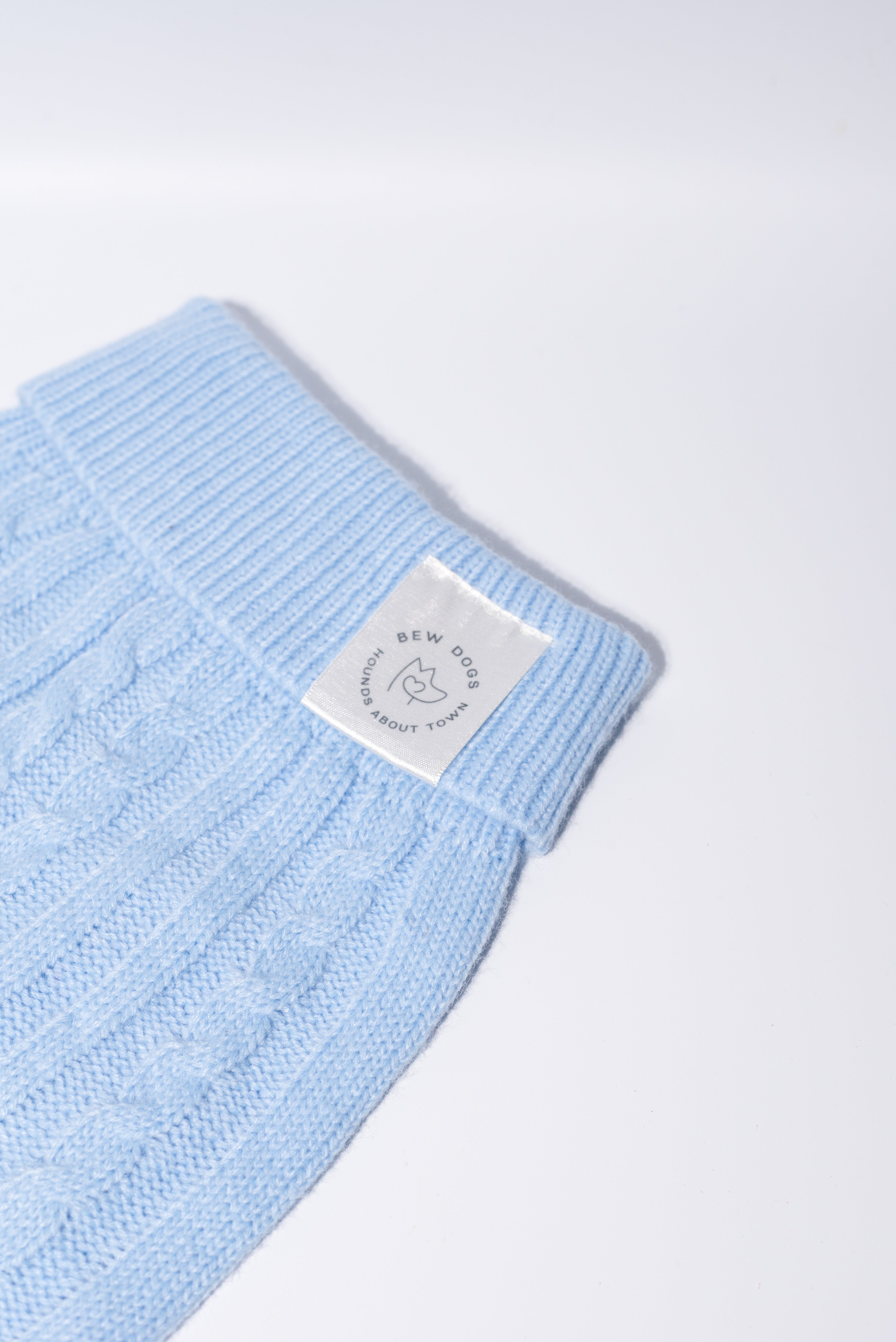 A close-up of a light blue cable-knit fabric dog jumper, featuring a white rectangular brand label that reads 'Bew DOGs’ - perfect for puppies and small dogs 