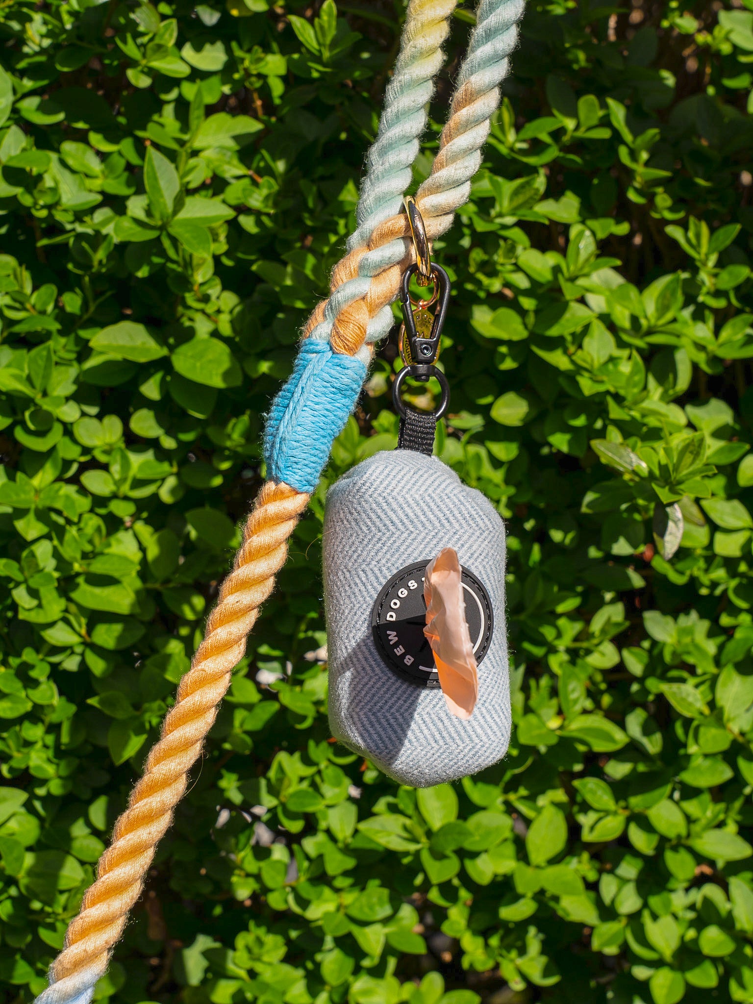 A soft blue poo bag holder, containing peach poo bags is shown in front of a leafy bush, hanging from a rainbow rope lead - demonstrating how this dog walking essential is used on everyday dog walks.