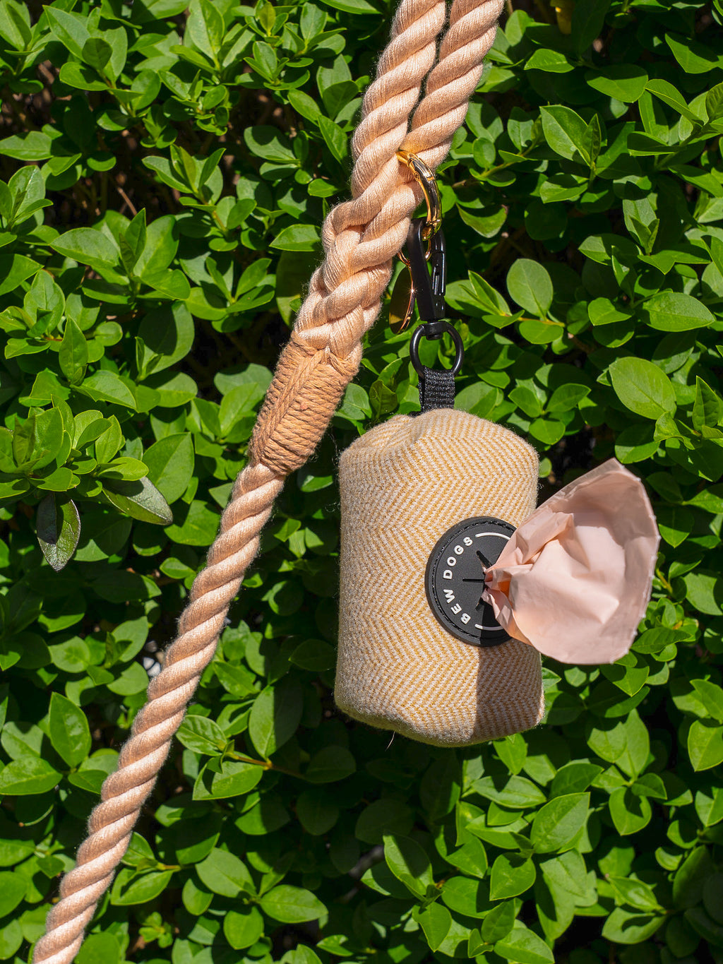 cream poo bag holder, containing peach poo bags is shown in front of a leafy bush, hanging from a peach rope lead - demonstrating how this dog walking essential is used on everyday dog walks.