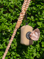 cream poo bag holder, containing peach poo bags is shown in front of a leafy bush, hanging from a peach rope lead - demonstrating how this dog walking essential is used on everyday dog walks.