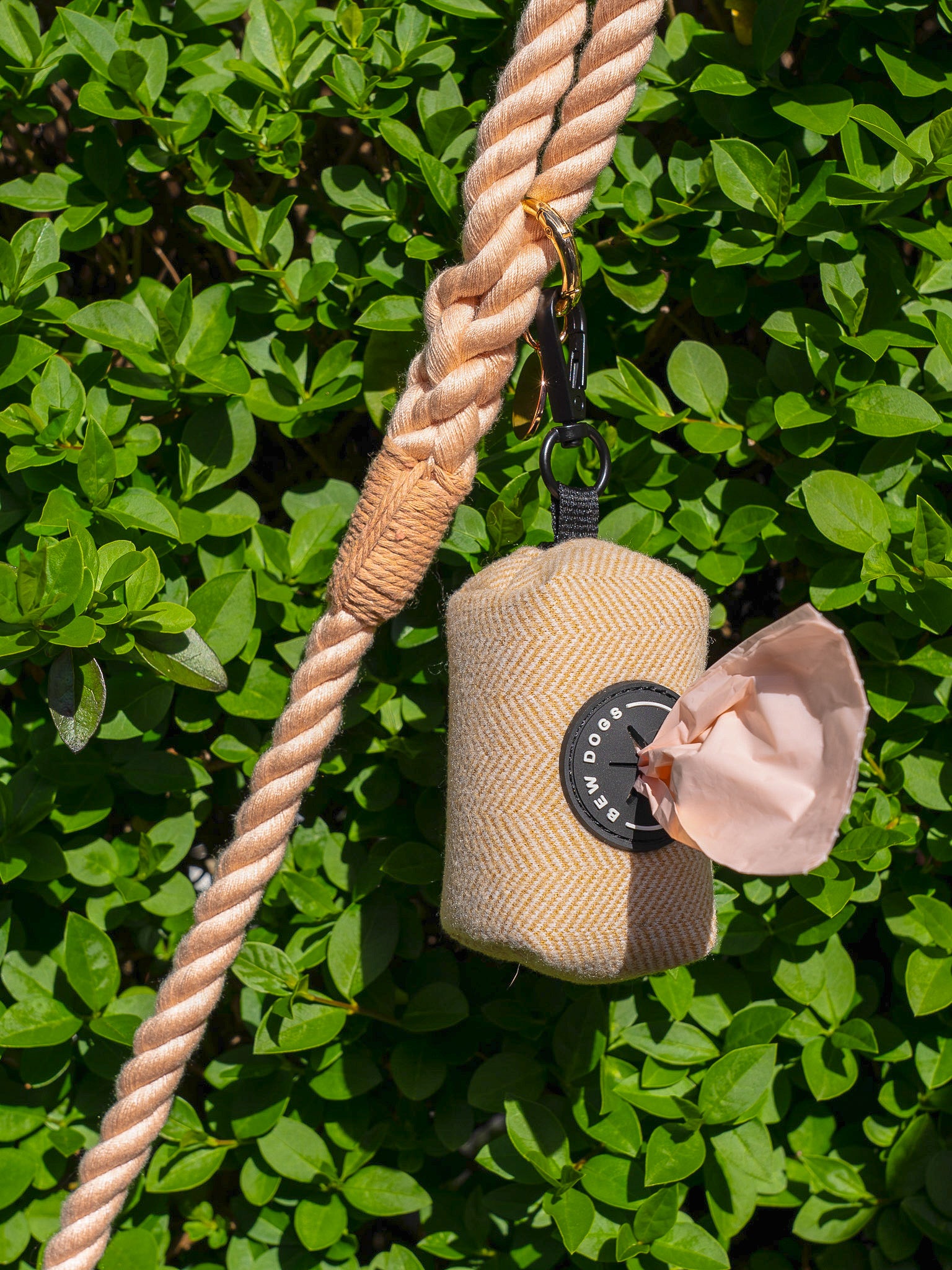 cream poo bag holder, containing peach poo bags is shown in front of a leafy bush, hanging from a peach rope lead - demonstrating how this dog walking essential is used on everyday dog walks.