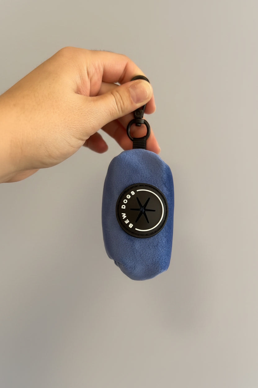 A deep blue velvet holder for poo bags, essential for all dog walks, is held against a grey studio wall. by Bew Dogs