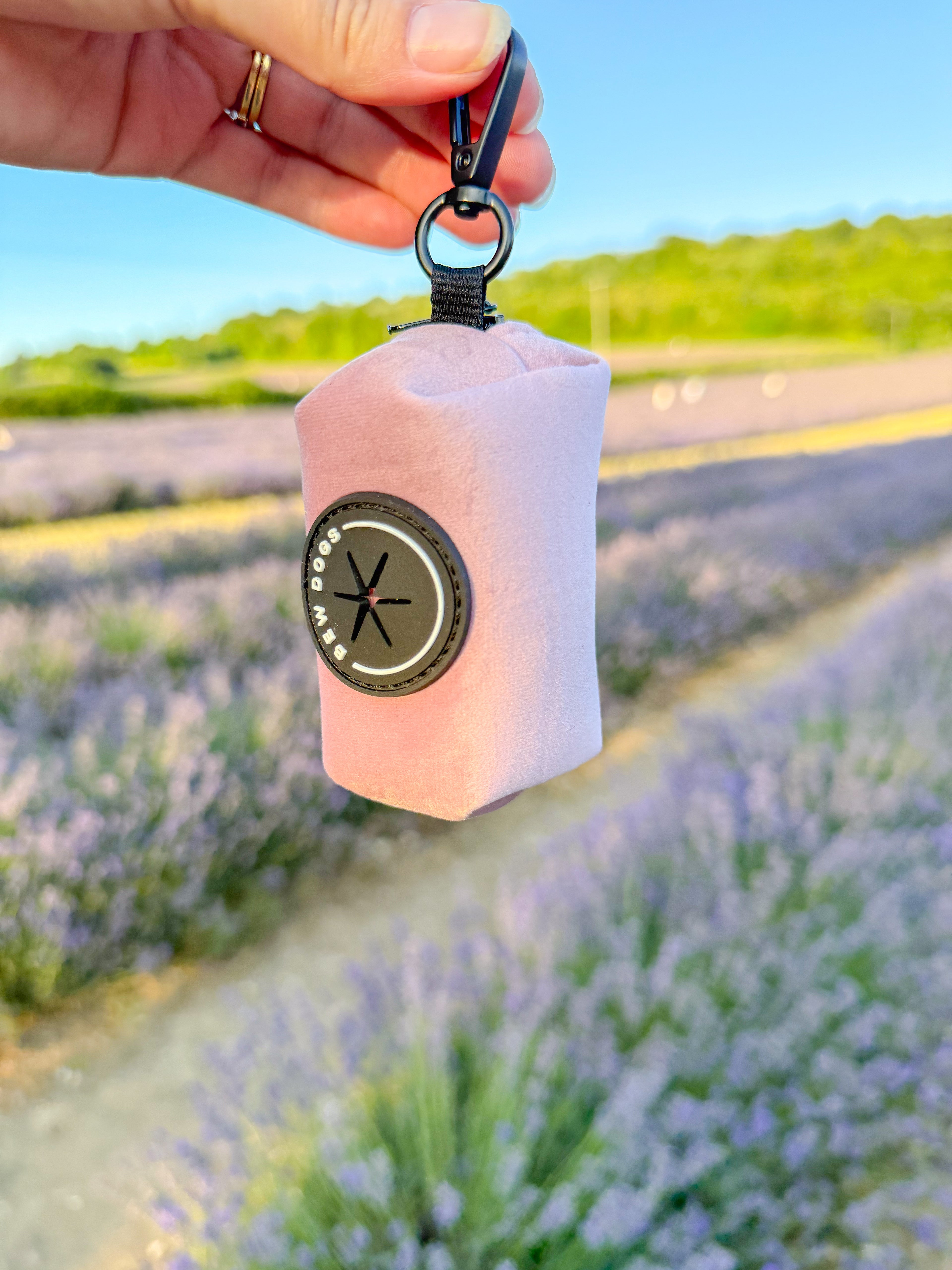 A dog walking essential pink velvet poo bag holder held by a hand, in the background is unfocused lavender fields 