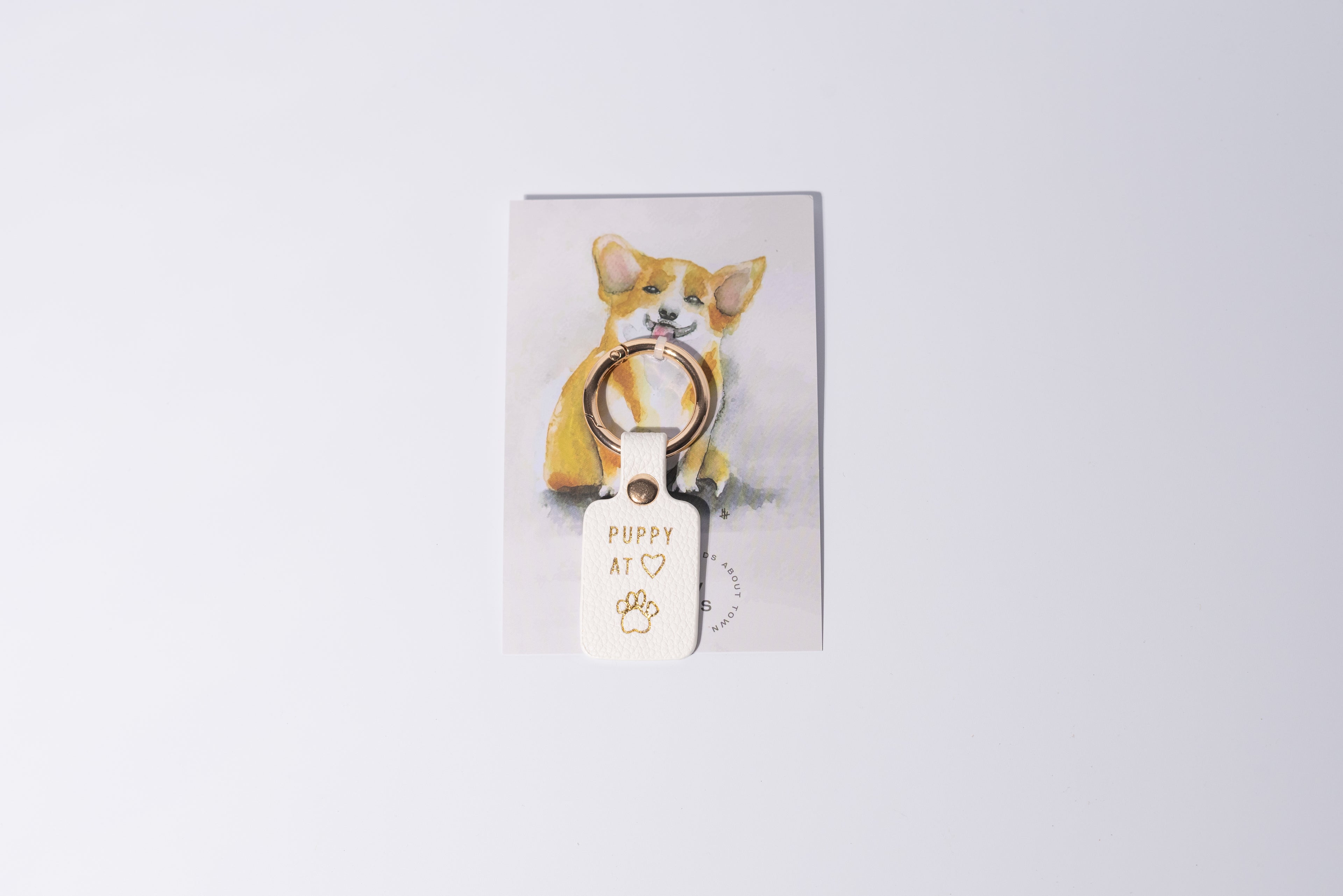 a white faux leather collar tag saying "puppy at heart" is connected to a promo card with a cute dog on it, the item is shown against a grey studio background