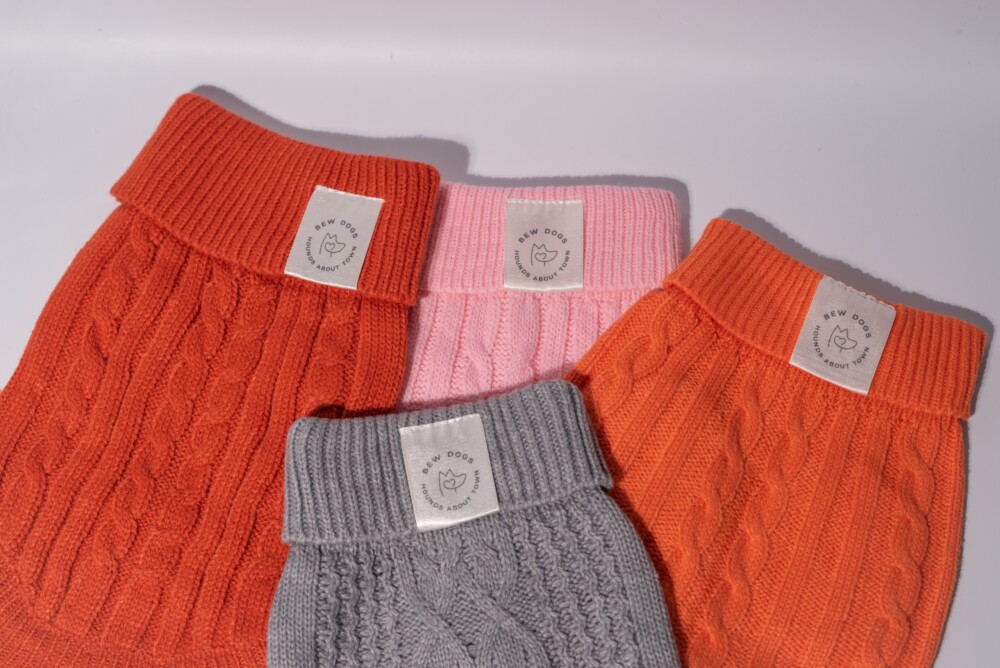 Four cable knit jumpers in orange, grey and pink, laid out against a white background