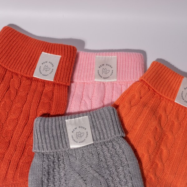 Four cable knit jumpers in orange, grey and pink, laid out against a white background