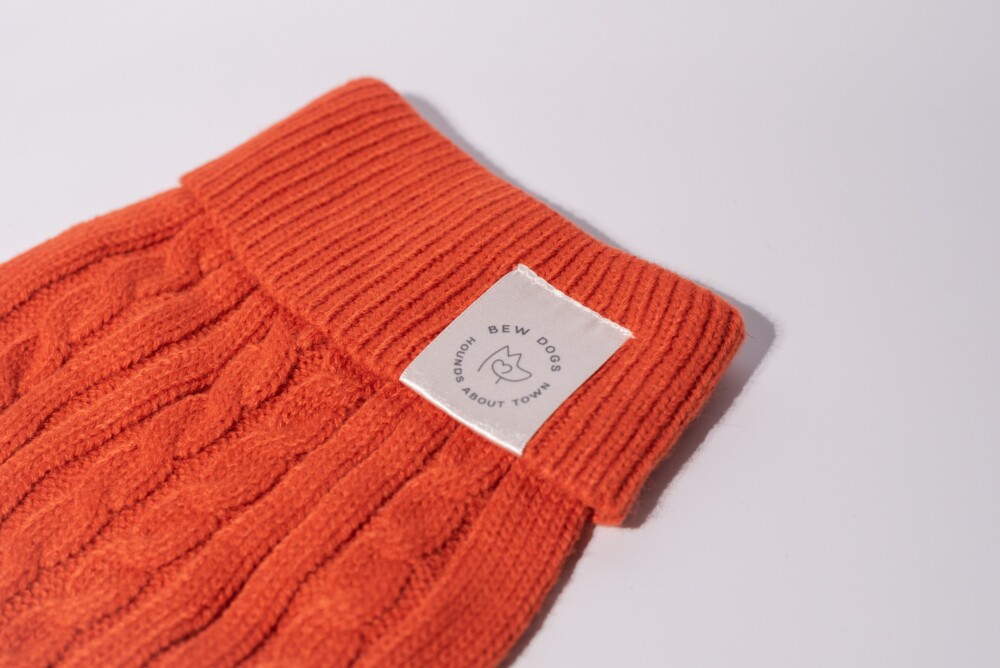 A close up of a burnt orange cable knit jumper on a white background