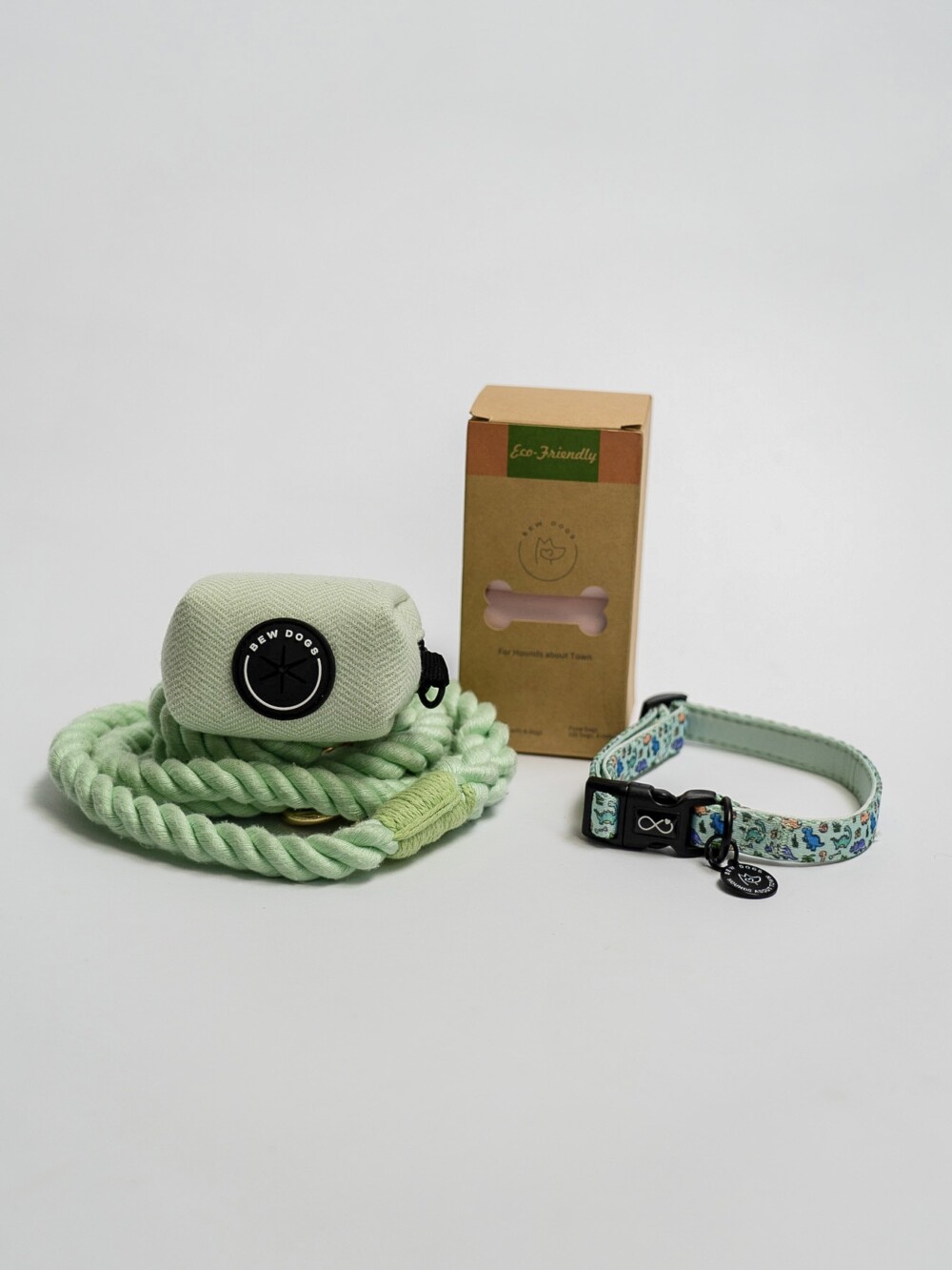 A mint green rope lead, herringbone poop bag holder, a box of eco-friendly poop bags and a blue/green collar, against a white background.