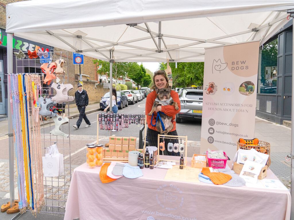 A market stall at a dog friendly event with lots of dog accessories, including colourful rope leads, tennis balls long lasting, hard wearing harnesses. with a banner and a woman holding a french bulldog.