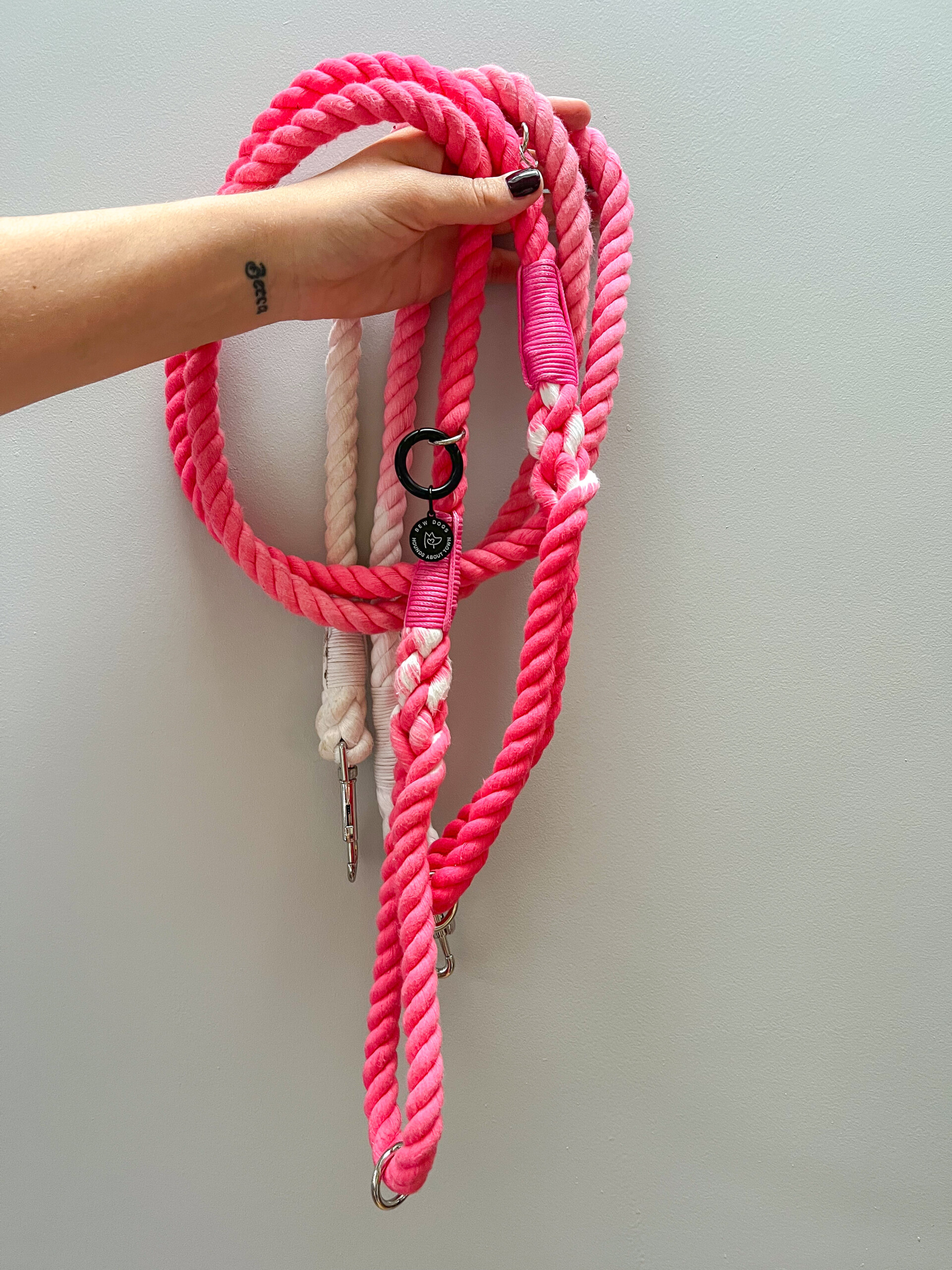 A pink ombre luxury rope lead, being held by a woman with deep and dark red painted nails against a grey painted wall