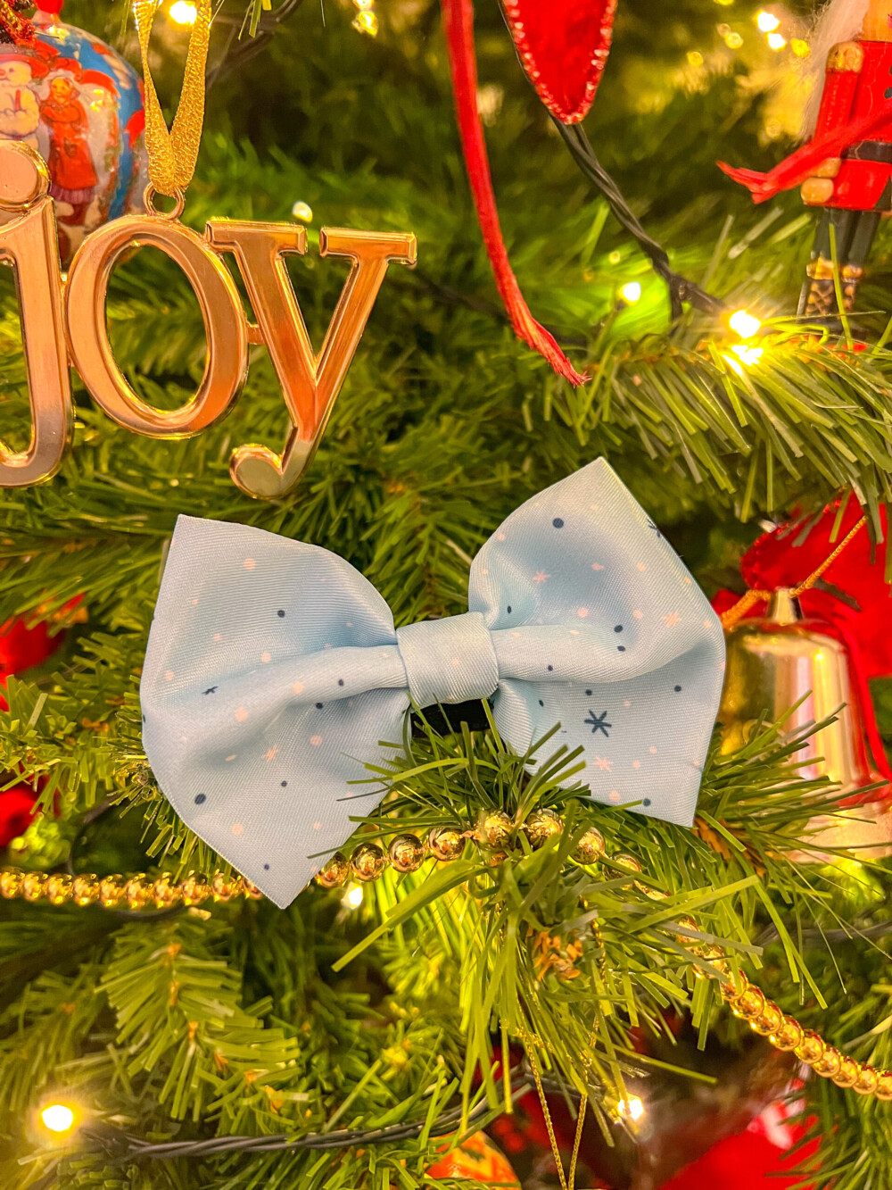 A winter dog bow tie, showing white and blue spots against a pale blue background, placed on a Christmas tree amongst decorations.