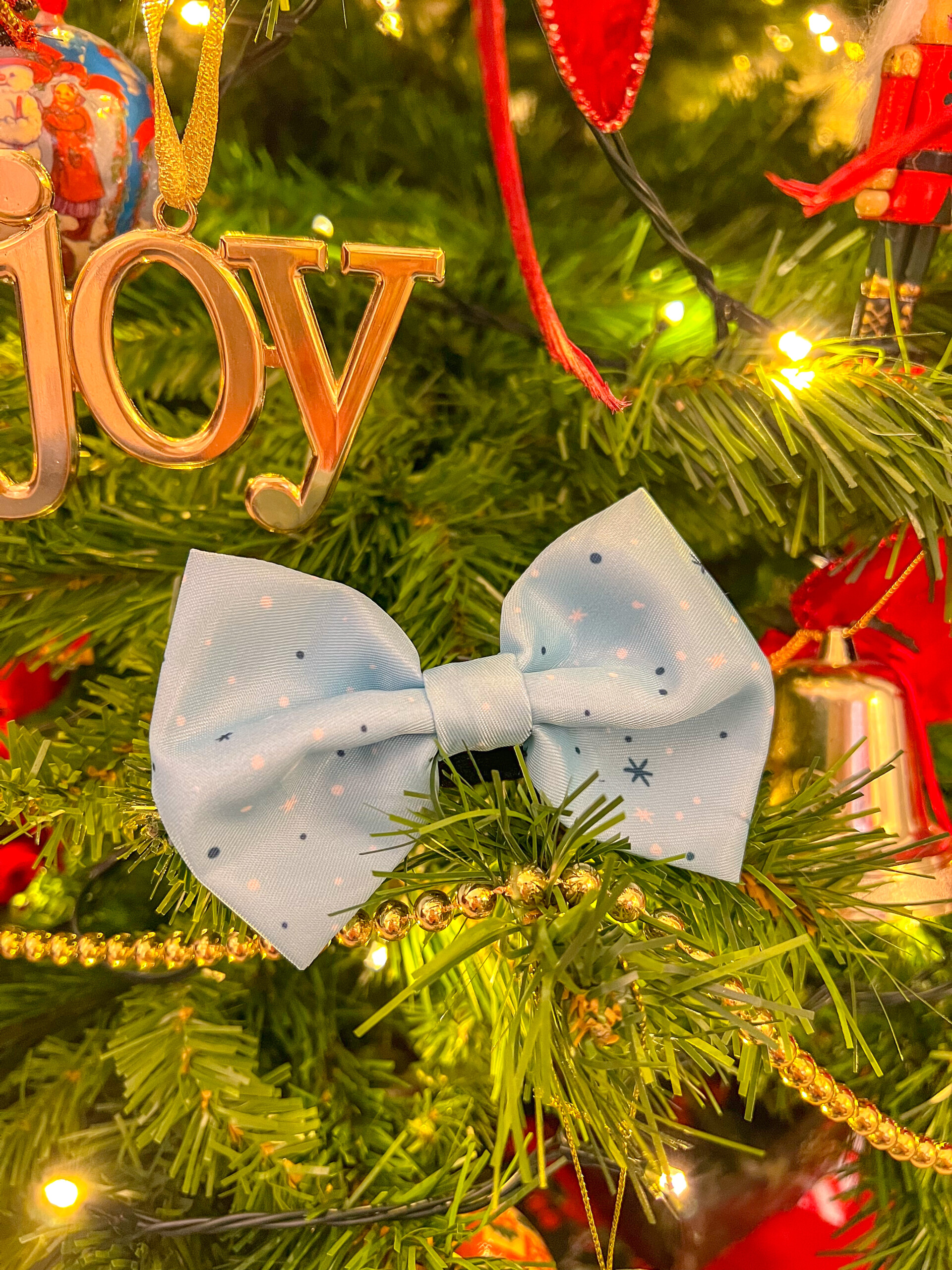 A winter dog bow tie, showing white and blue spots against a pale blue background, placed on a Christmas tree amongst decorations.