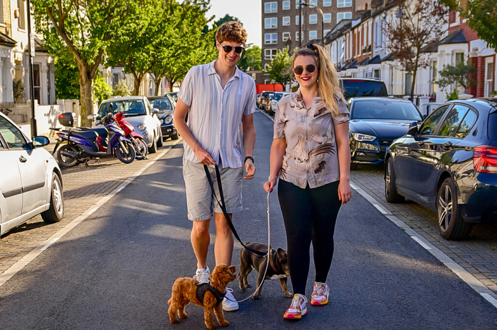 a young couple are smiling and walking down a london street outside their doggy day care in fulham, SW6 with two small dogs on leads