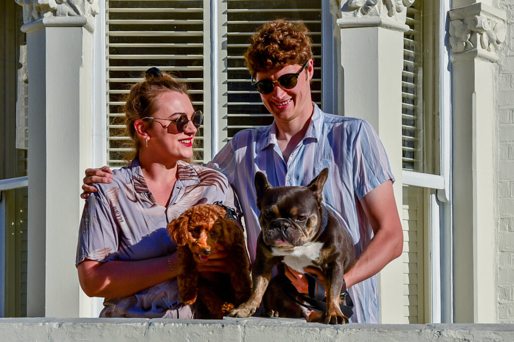 a young couple stand outside the best doggy day care in fulham for small and medium breed dogs, they are wearing sunglasses, it is a hot day and they're holding a poodle and french bulldog. 